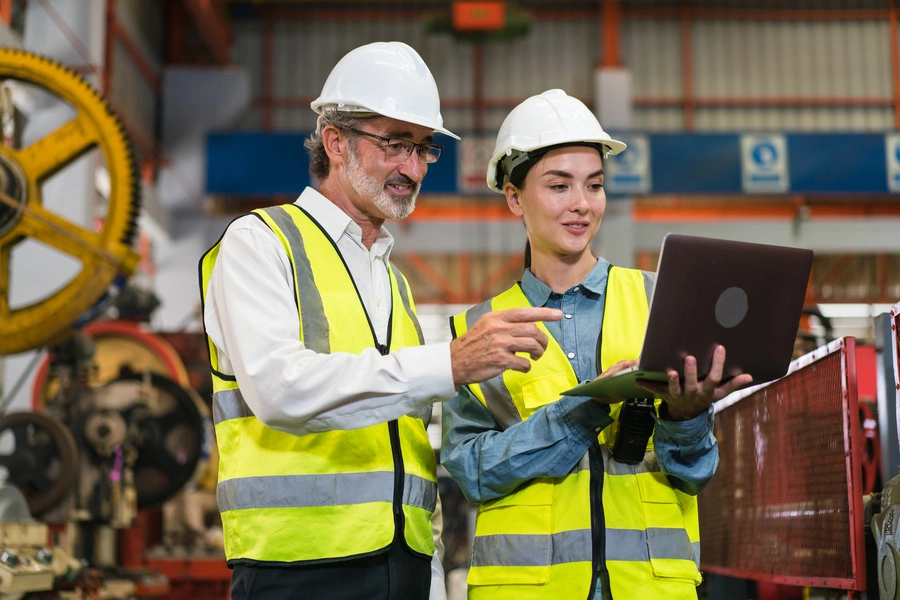 zarządzanie zmianą (kobieta i mężczyzna w kamizelkach odblaskowych stoją na hali, kobieta pokazuje coś na laptopie)